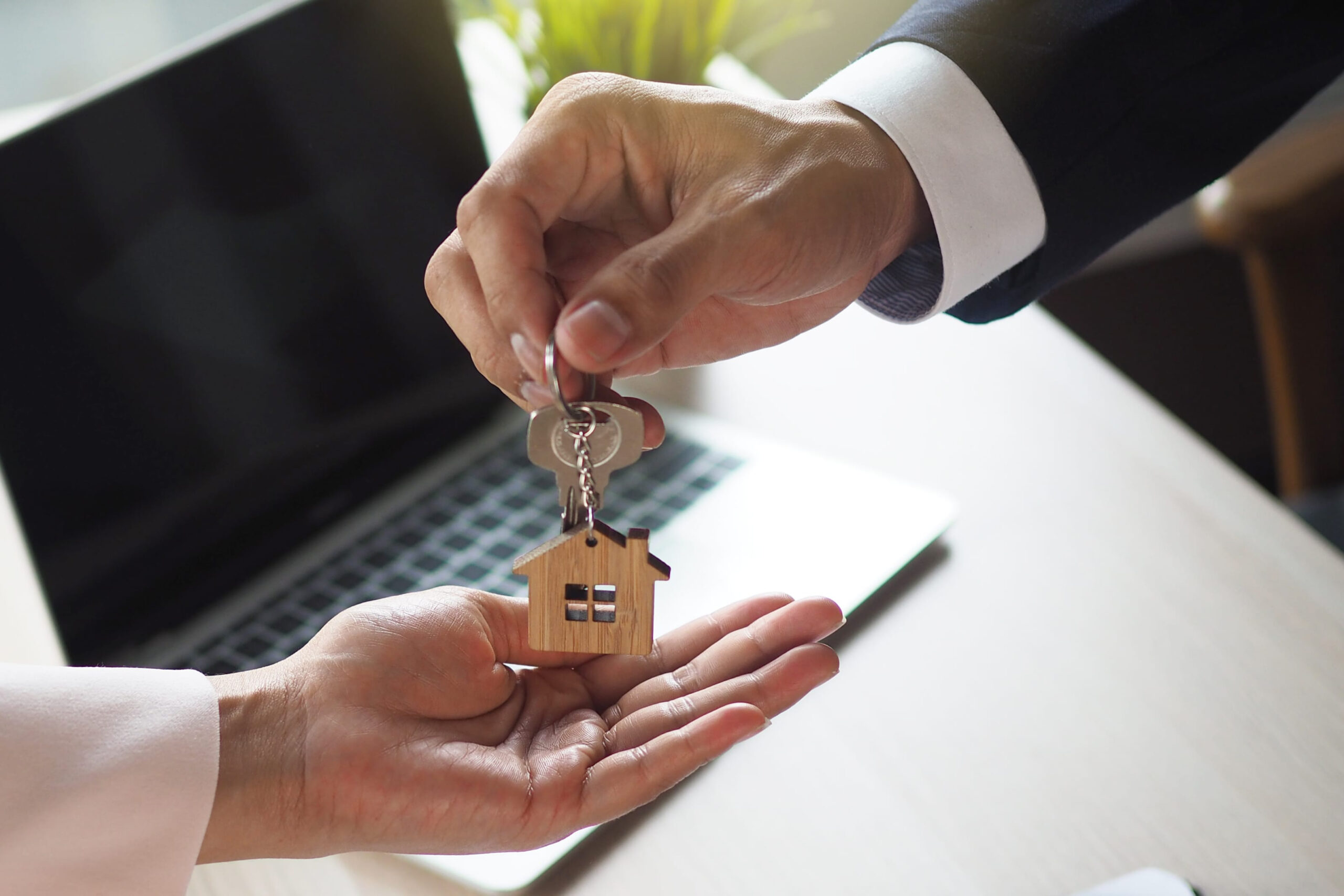 A real estate professional handing over house keys with a wooden home keychain to a new homeowner during a property transaction, symbolizing a successful home sale or cash home purchase.